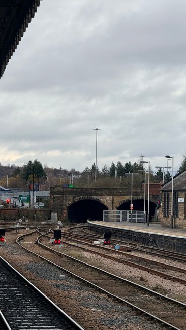 The image shows an empty railway platform under cloudy skies, with multiple train tracks curving towards a stone tunnel in the distance. The platform is lined with modern lampposts and safety barriers