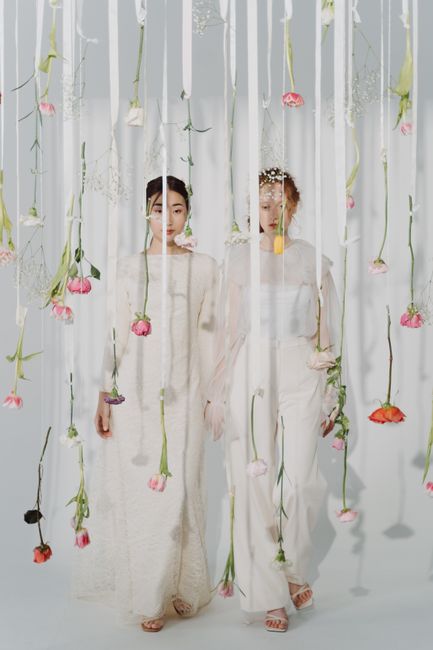Two women dressed in elegant white outfits stand behind a delicate curtain of hanging pink and peach roses with green stems and leaves, creating a soft, floral backdrop. The women are positioned side