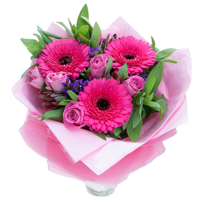 I see this bouquet as a balance of exuberance and softness: three bold gerbera daisies in a saturated fuchsia occupy the visual centre, their ring of petals surrounding dark, almost black centres that give the blooms a theatrical clarity. Interspersed are roses in a softer lavender-tinged pink, each one still holding the intimacy of a bud so the arrangement reads as both fresh and reserved. Sprays of tiny purple statice weave through the composition, adding a whisper of wildflower charm and a pleasing textural counterpoint to the larger, smooth petals. Deep green foliage - broad leaves like salal alongside longer, bluish eucalyptus stems - forms a generous backdrop that intensifies the pink notes and brings a gardenlike freshness to the whole. The bouquet has been wrapped with intent: an inner layer of soft pale-pink tissue cushions the stems while an outer translucent fuchsia-pink cellophane, with a subtle crinkle, reflects light and ties the colour story together. The bottom edge of a clear glass vase is visible, signalling that the arrangement is ready to be placed on a side table or bedside unit. In diffused daylight the colours read true and luminous; the roses give off a gentle, classic scent while the greenery adds a faint dewy, almost citrus-green aroma. Ideal for an anniversary, a birthday, or a surprise 'just because' delivery, this piece would be well suited to brighten a home in Cranham village or to sit as a welcoming display in a local tea room, crafted with the hands-on care of an experienced florist.